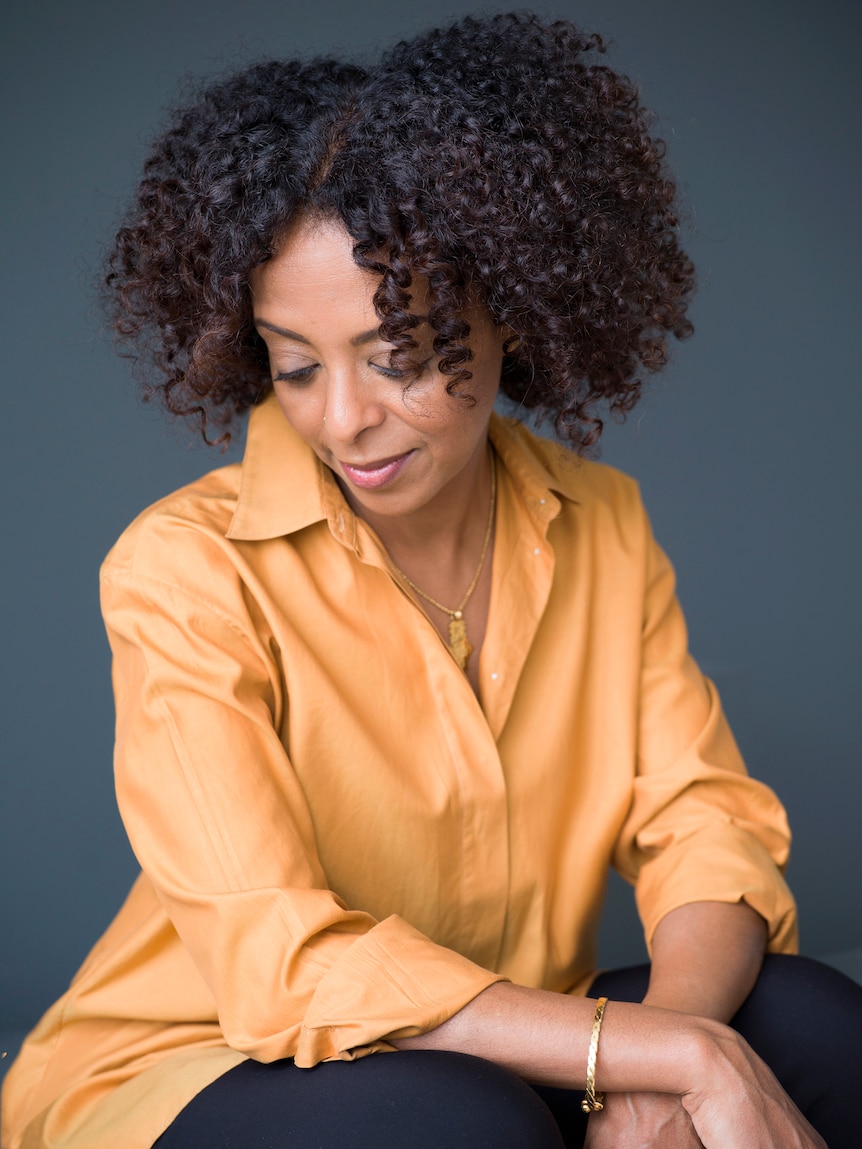 Portrait of a middle-aged Ethiopian born woman with curly hair and a warm orange collared top. She's looking downwards 