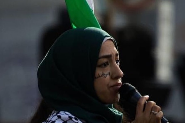 Aseel wearing a black hijab, holding a microphone with a Palestinian flag behind her.