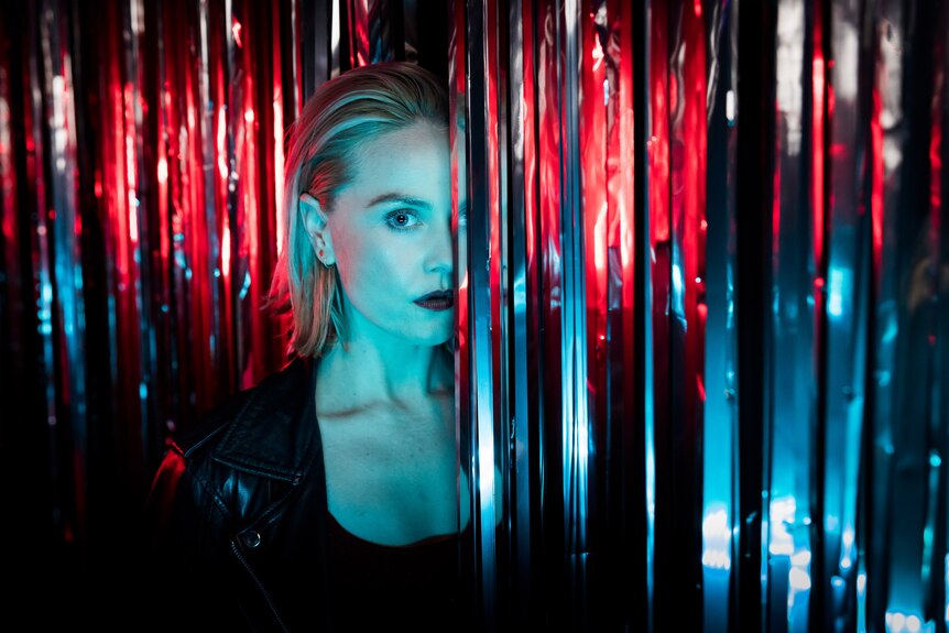 White woman with blonde lob wears black singlet and leather jacket and stands, lit in blue and red, between silver streamers.