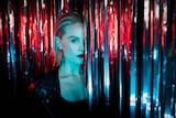 White woman with blonde lob wears black singlet and leather jacket and stands, lit in blue and red, between silver streamers.