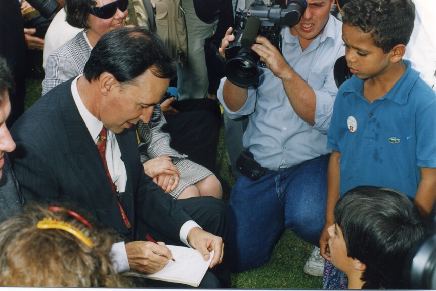 Paul Keating signs autographs for Aboriginal children, surrounded by photographers.