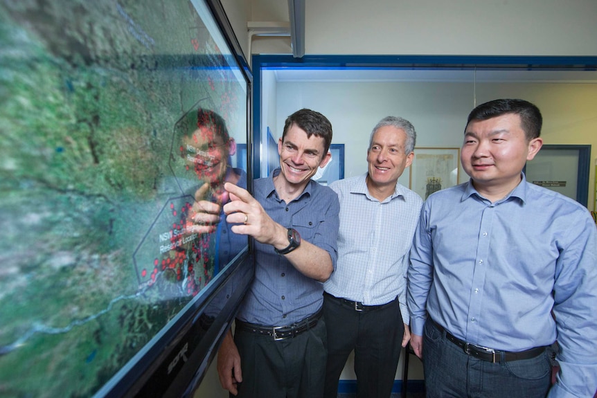 Three men stand in a row and point at a map.  The middle one has white hair and brown eyes.