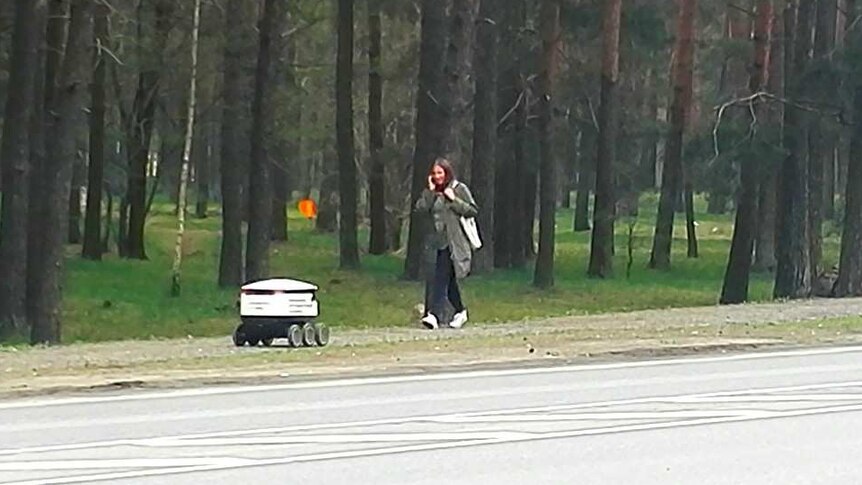 A small delivery bot drives down the footpath as a woman walks past.