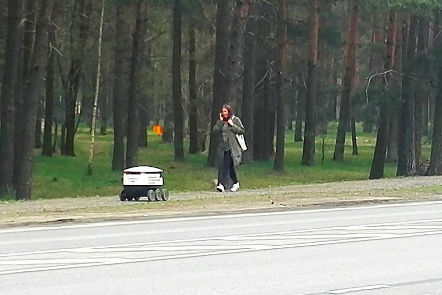 A small delivery bot drives down the footpath as a woman walks past.