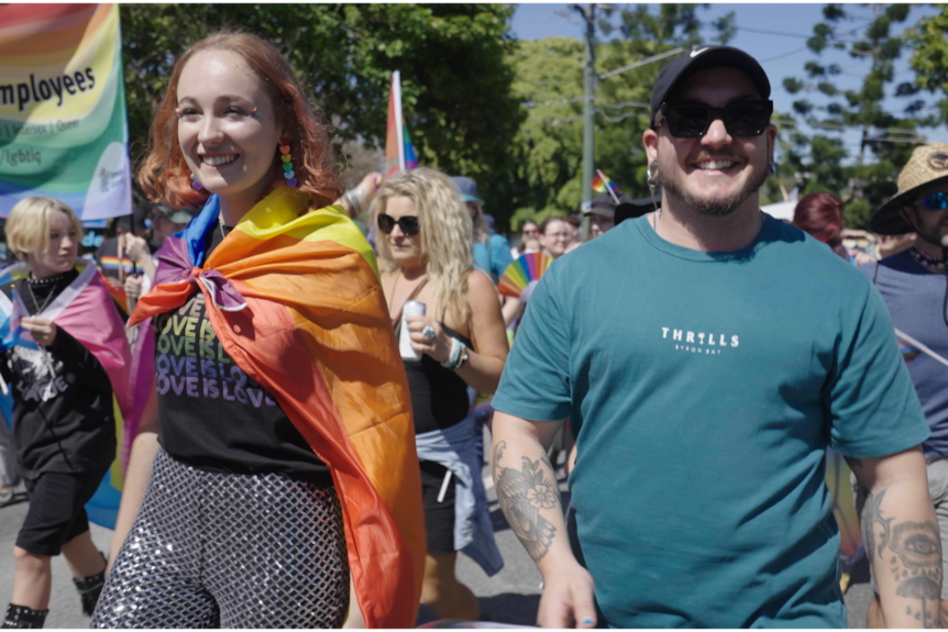 A man and a woman march next to each other in a pride parade. The woman has a rainbow flag over her shoulders