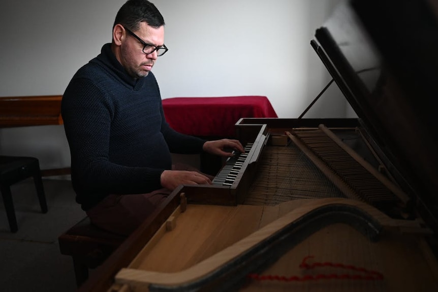 A man in navy jumper and thick black-rimmed glasses playing a heritage piano.