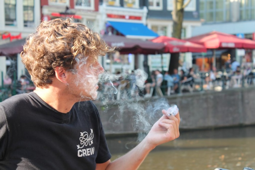 A man with blond curly hair holds a joint while exhaling a cloud of smoke, behind him a canal and some shops.