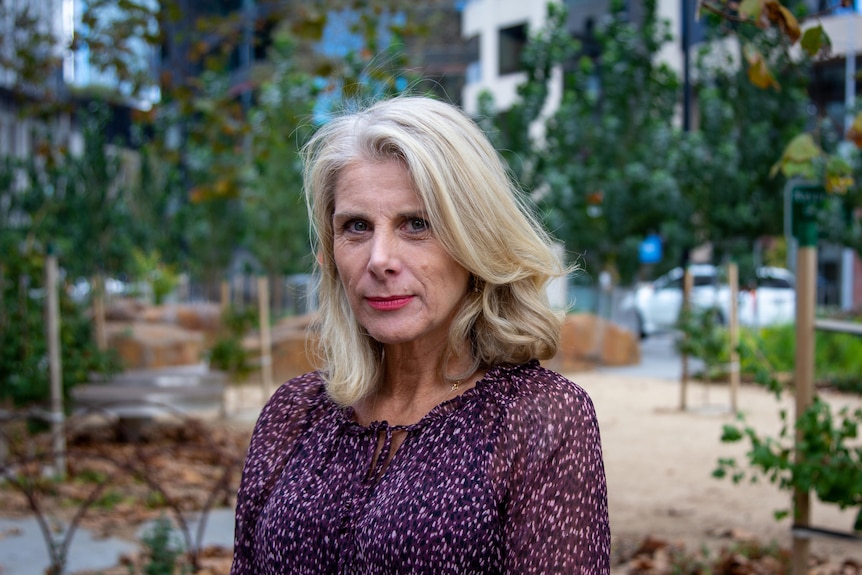 A middle aged woman with blonde hair, purple top, red lipstick stares into the camera in front of landscaped tree area