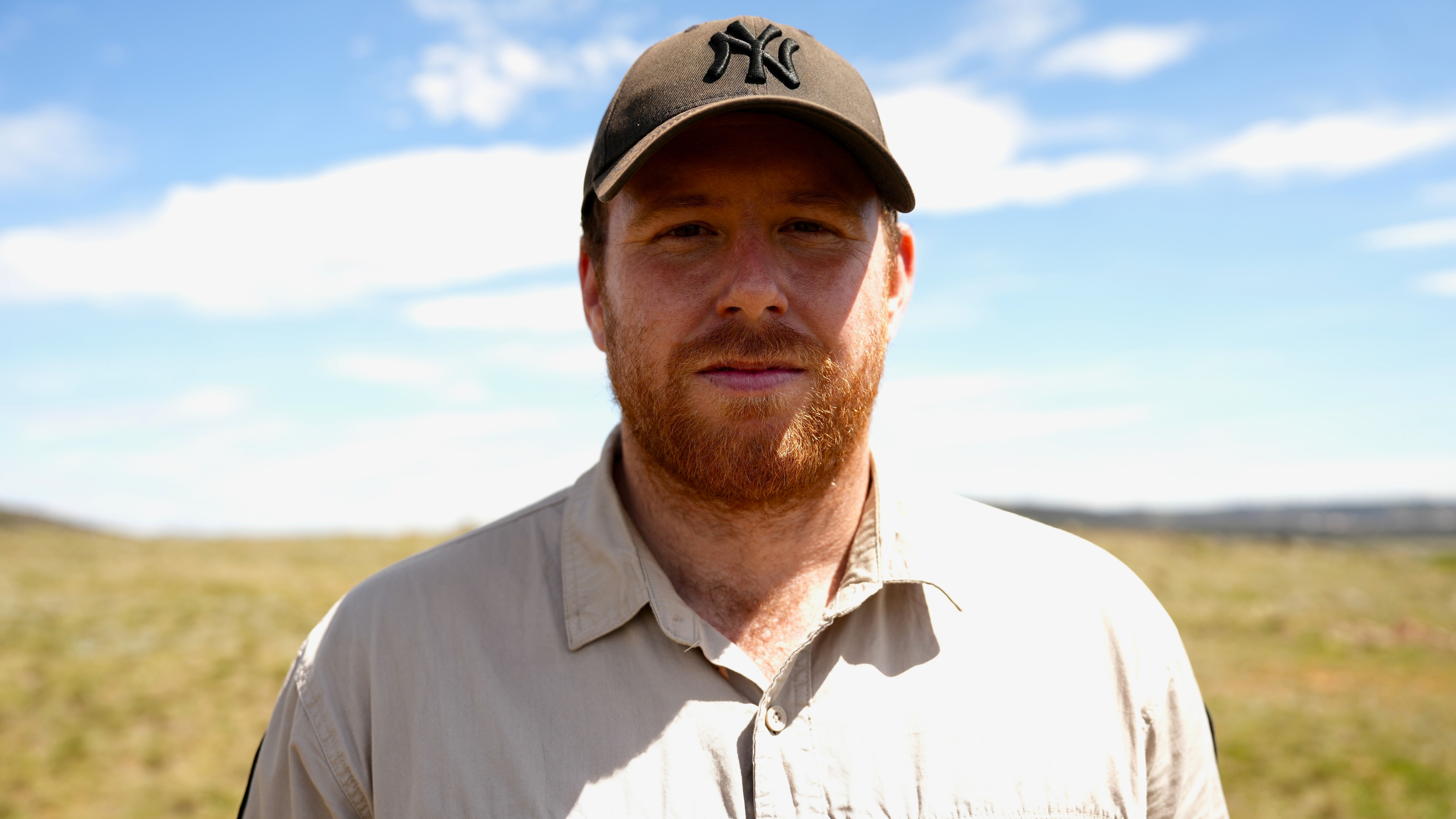 A man wearing a beige button-down shirt and black cap stands in a paddock smiling lightly.