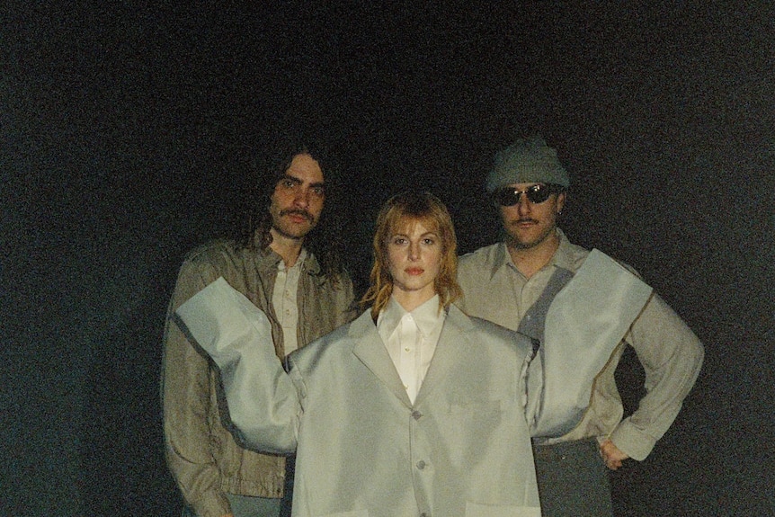 Three members of Paramore in a dark room. Hayley Williams wears an oversized jacket like Talking Heads' David Byrne