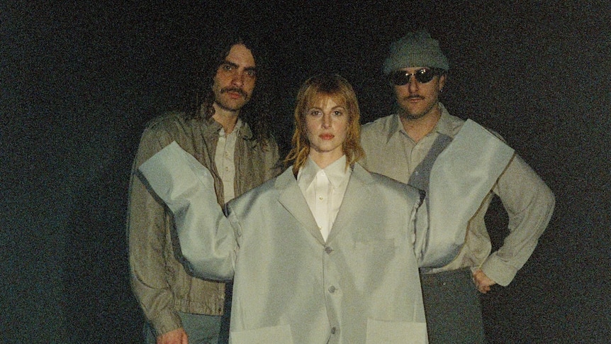 Three members of Paramore in a dark room. Hayley Williams wears an oversized jacket like Talking Heads' David Byrne