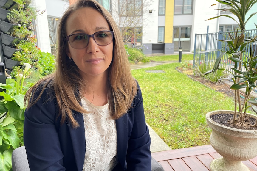 Woman sitting in a garden. She's wearing a white shirt and navy jacket.