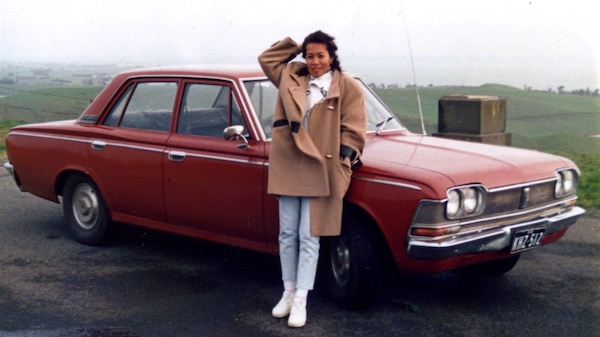 You view a young Filipina leaning against a vintage red sedan, as green rolling hills stretch out behind her to the horizon.