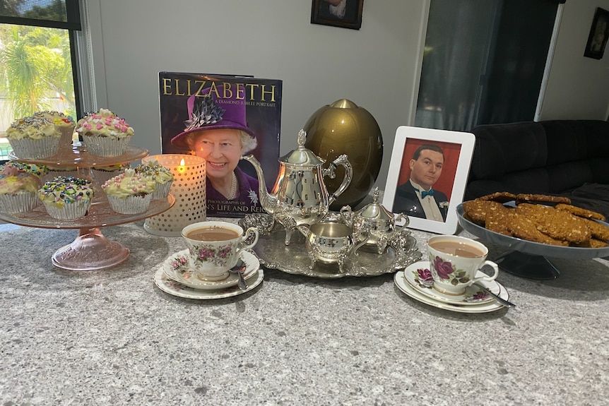 A morning tea laid out with tea cups and cupcakes. A book about Queen Elizabeth II is displayed with a photo of Barry.