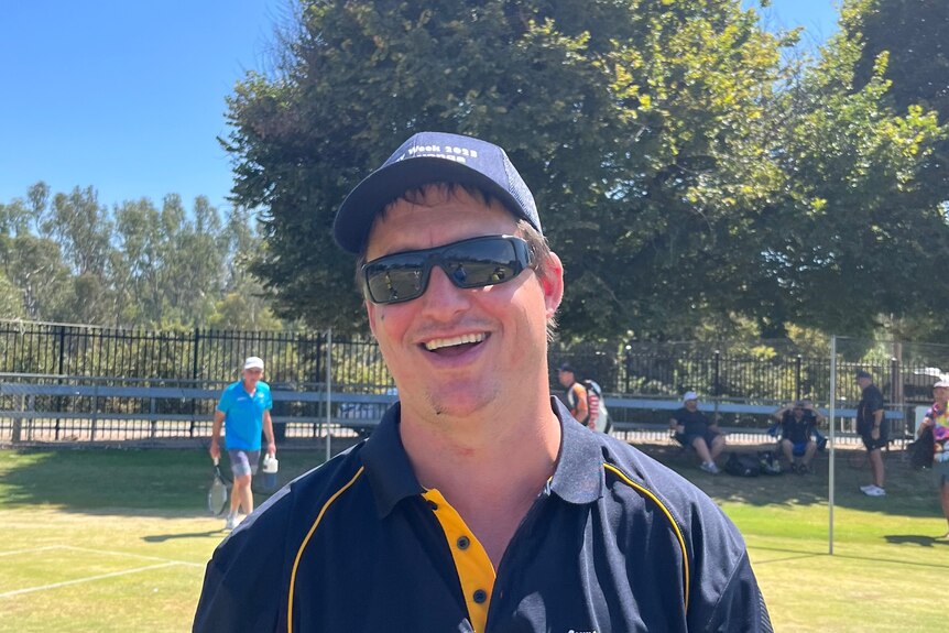 Man in navy cap and polo top and wearing dark sunglasses smiles 