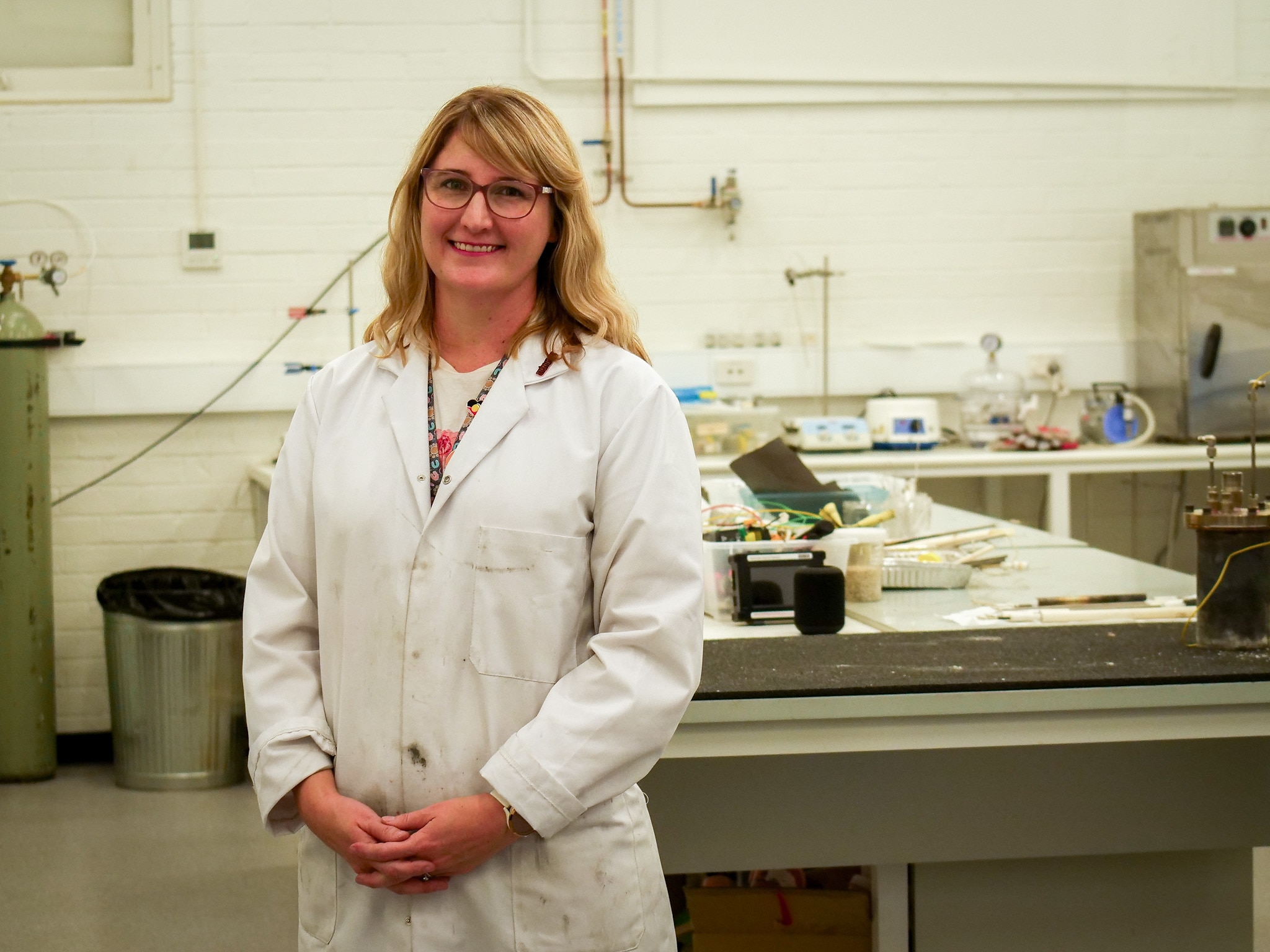 A blonde-haired woman in a white lab coats stands smiling to camera in a scientific lab.