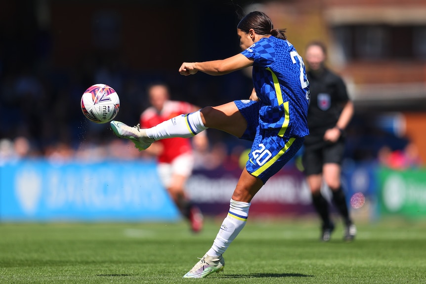 A female soccer player wearing blue and yellow kicks the ball in the air during a match