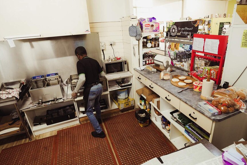 A man in jeans and a black apron stands behind a grill and fryer in a small kitchen.