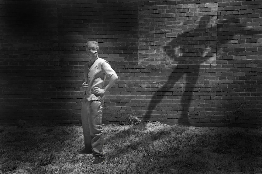 A black-and-white photo of man wearing surgical scrubs casting a shadow of a cape-wearing superhero.   