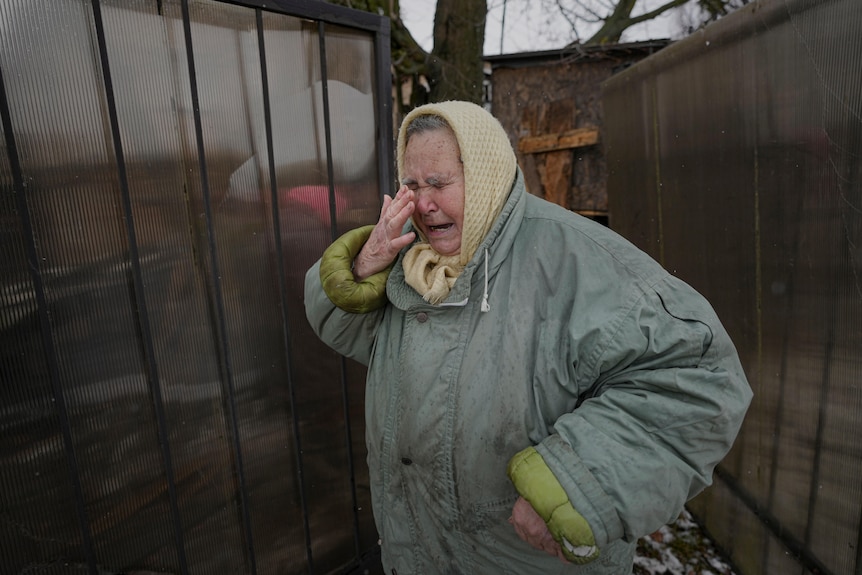 Elderly woman in winter clothes brings a hand to her eyes as she cries outside a damaged house.