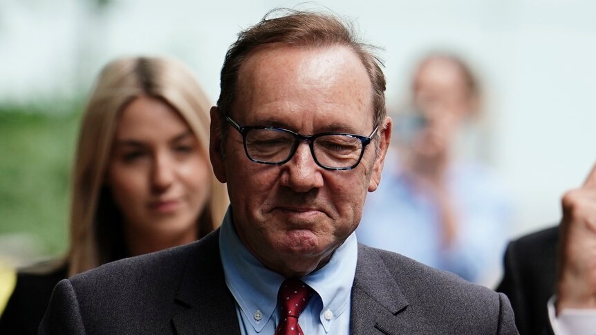 Kevin Spacey is pictured wearing glasses, a grey suit with a blue shirt and red tie. He looks down. Behind him a blonde woman.