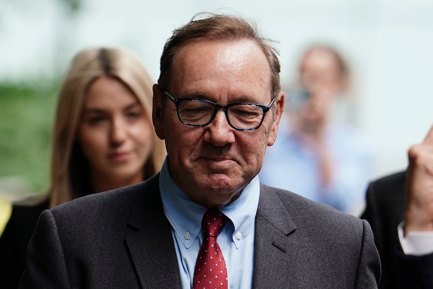 Kevin Spacey is pictured wearing glasses, a grey suit with a blue shirt and red tie. He looks down. Behind him a blonde woman.