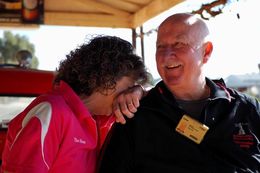 A middle aged heterosexual couple are laughing. The lady, with curly brown hair, buries her face in her husband's shoulder.