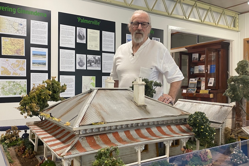A man with grey hair and a grey beard wearing glasses looks at a model of a homestead 