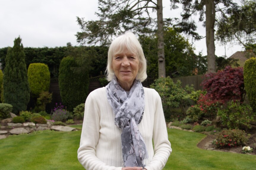 A woman with white hair and wearing a scarf and white shirt smiles in a garden.