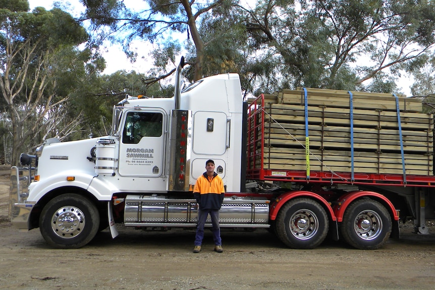 A white semi-trailer attached to a trailer carrying wooden beams is parked with its side to the camera, with a worker in front.