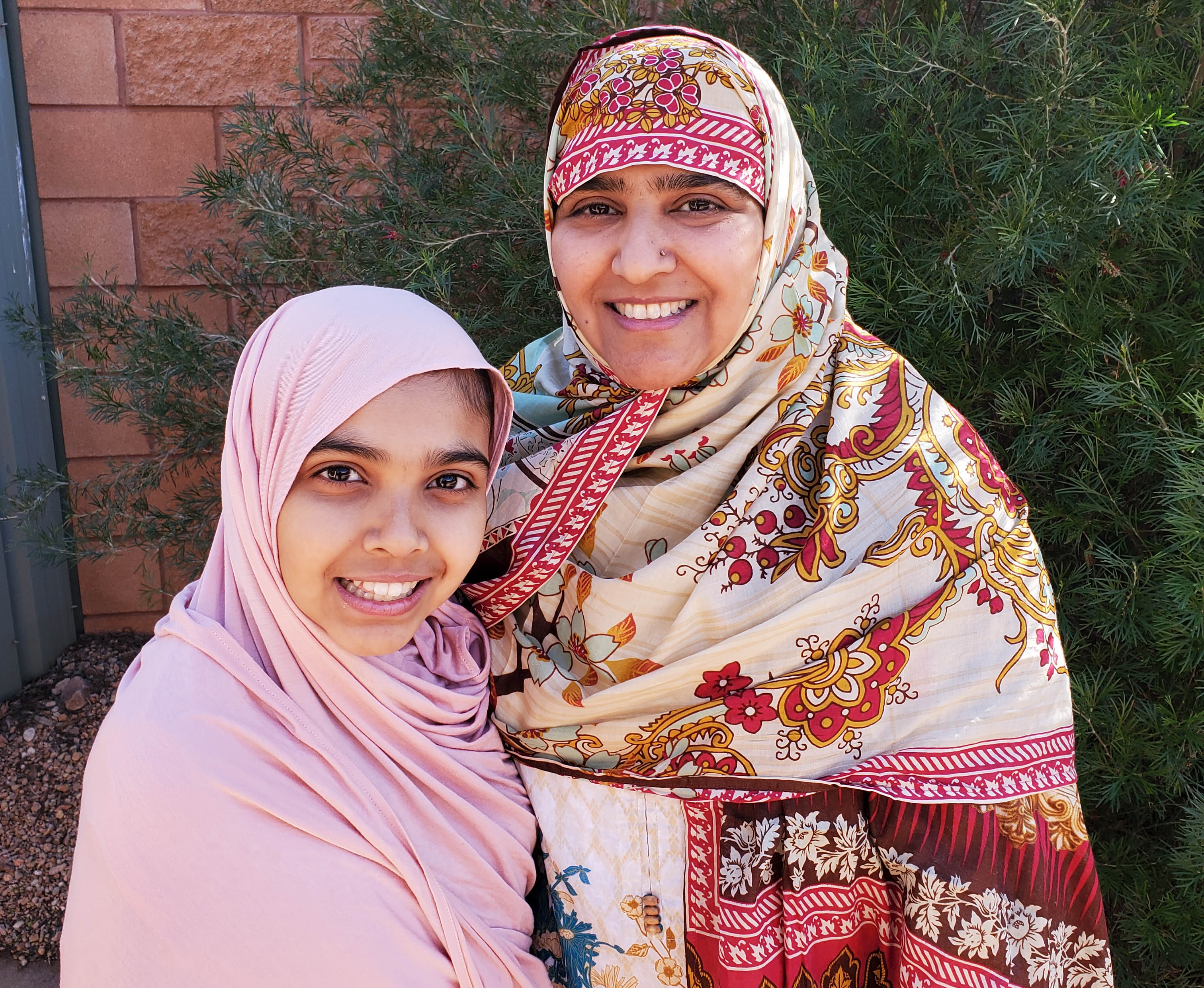 A woman smiling with a patterned hijab with her young teenage daughter smiling and wearing a pink hijab.