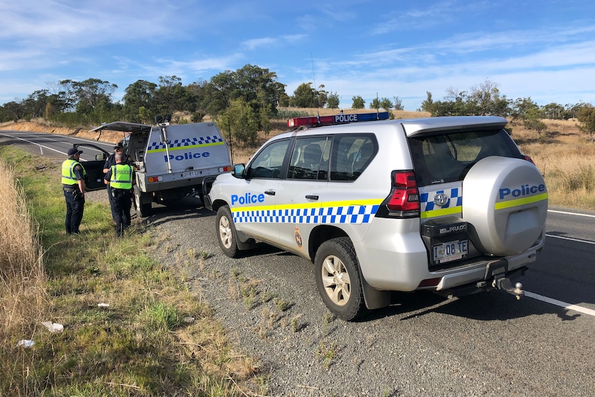 Two police officers stand near two police cars on the side of a road.