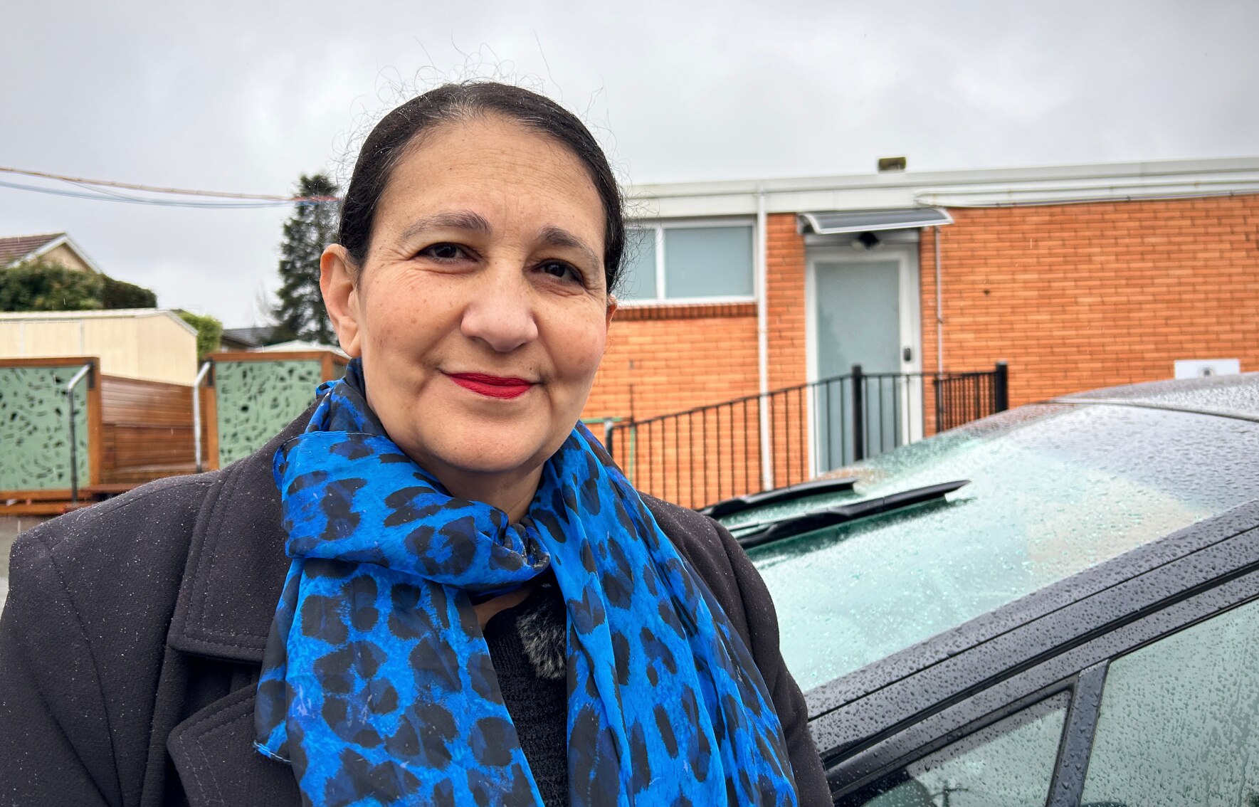 A woman stands outside a building and a car and smiles.
