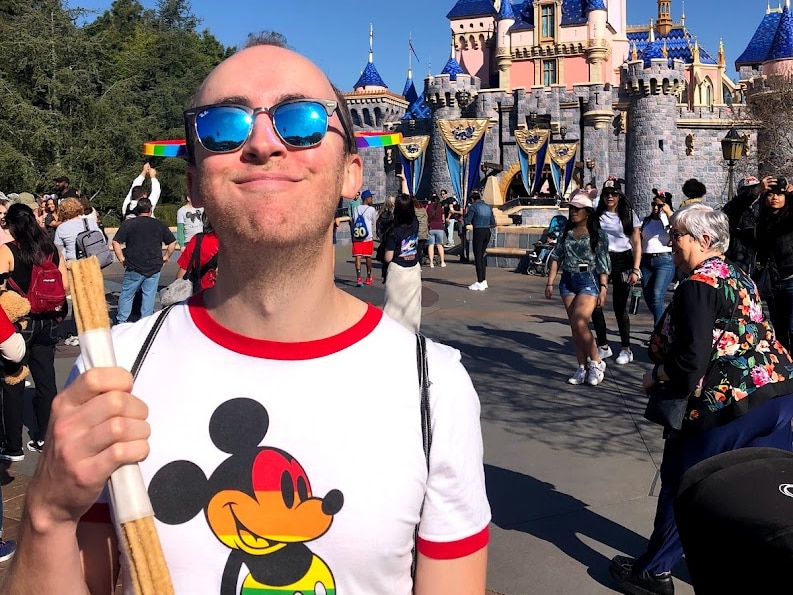 A happy Joel Callen in front of a Disneyland castle wearing sunglasses and a rainbow Mickey shirt, holding a churro.