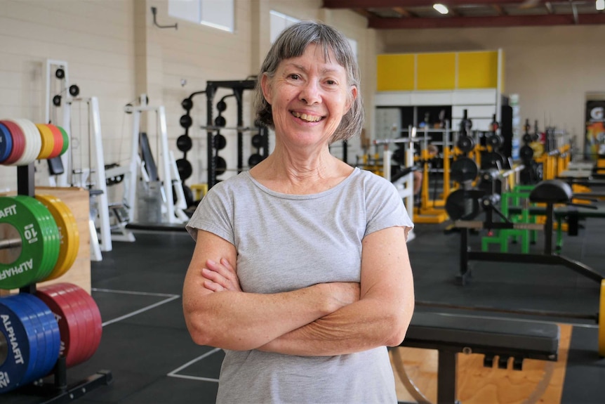 Smiling woman with grey hair with arms crossed stands in a gym.