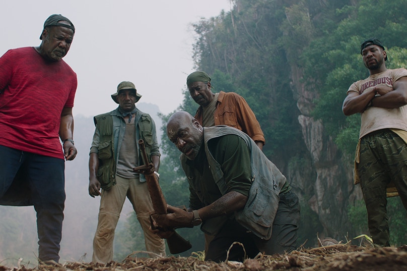 On hazy day in mountainous jungle four men in camping attire stand above and around a man kneeling in dirt ditch holding rifle.