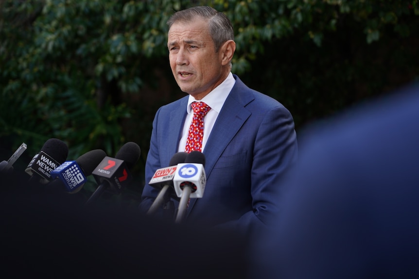 Roger Cook wears a blue suit and red tie, standing at microphones talking to the media.