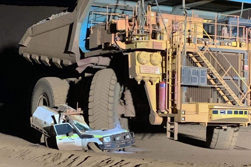 A white four wheel drive ute is crushed beneath the tyre of a large yellow dump truck