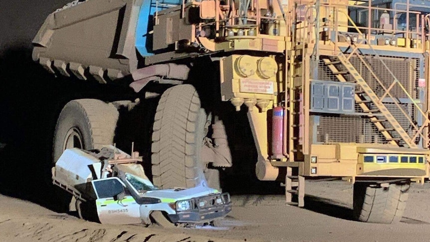 A white four wheel drive ute is crushed beneath the tyre of a large yellow dump truck