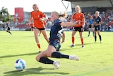 A woman soccer player tries to get the ball as it is rolling over the line as other players in orange shirts watch on