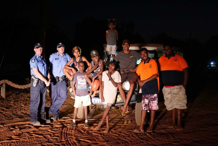 Two police officers and a group of Indigenous children standing in front of a Land Cruiser at night.