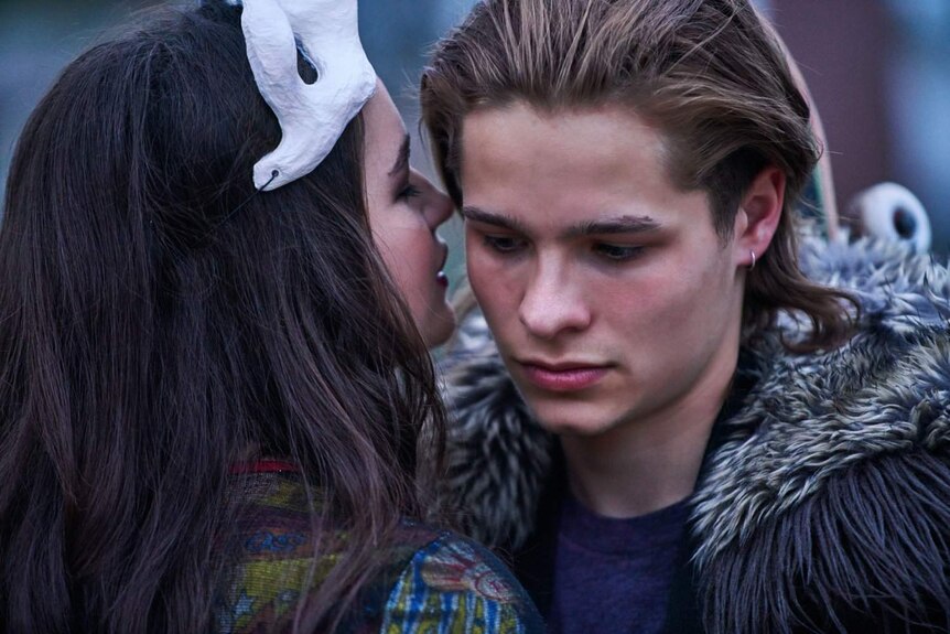 A teenage girl wearing a mask whispers into the ear of a teenage boy.