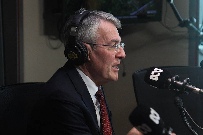 A man with grey hair, glasses and a dark suit sits with headphones on and speaks into a microphone. 