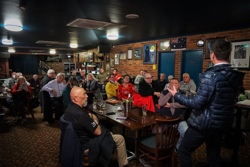 Man in black puffer jacket speaks to a crowd of people inside a brick pub.