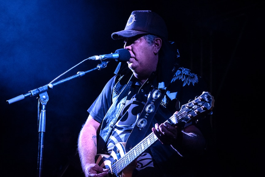A man in the dark playing his guitar while singing with a blue tinge stage light.