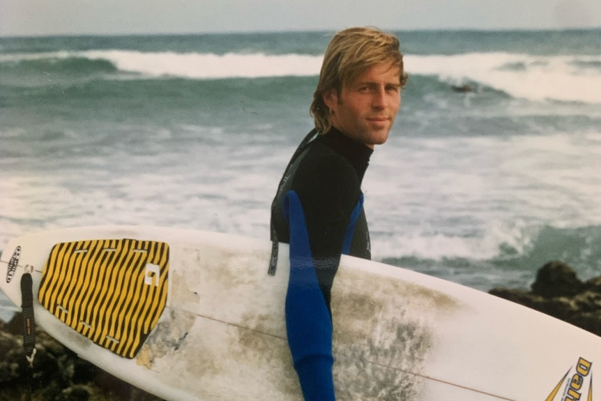 An old photo of a smiling man with long blonde hair wearing a wetsuit and carrying a surfboard.