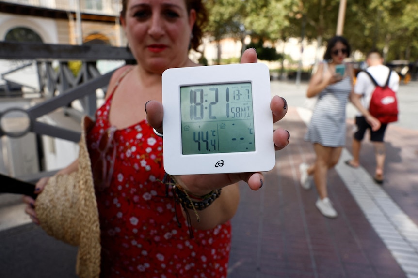 A woman holds a digital thermometer facing the camera and displays a temperature of 44 degrees Celsius. 