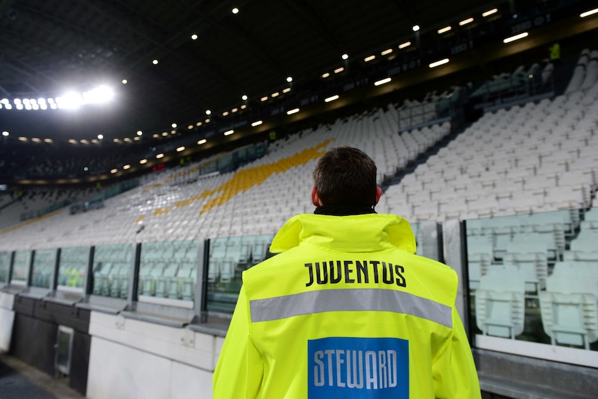 Juventus is written on the back of a steward's high-vis jacket as he looks at empty seats in a stadium.