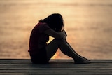 Girl sits alone with her head down in silhouette near a body of water.