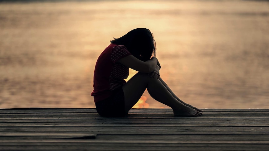 Girl sits alone with her head down in silhouette near a body of water.